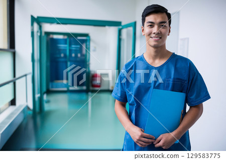 Asian man wearing blue scrubs standing in hospital corridor holding blue folder, copy space Asian man wearing blue scrubs standing in hospital corridor holding blue folder, copy space 129583775