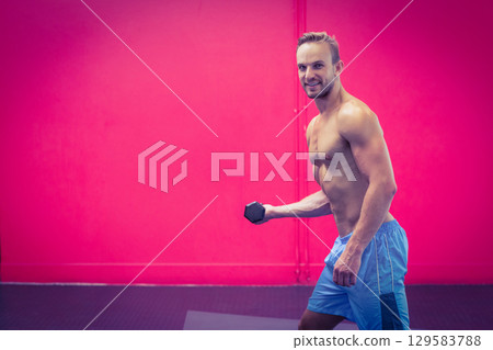 Muscular man holding hex dumbbell in pink studio beside vertical pipe and rubber mat, copy space 129583788