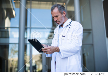 Senior male physician standing at clinic door reviewing tablet and wearing stethoscope, copy space 129583795