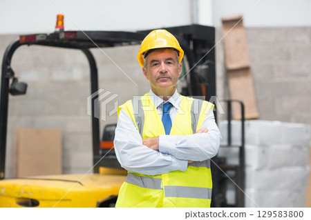Middle aged supervisor wearing yellow hard hat and safety vest standing by forklift in warehouse Middle aged supervisor wearing yellow hard hat and safety vest standing by forklift in warehouse 129583800