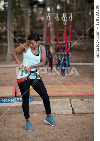 Woman fastening safety harness with carabiners around waist on wooden platform at park ropes course 129583806