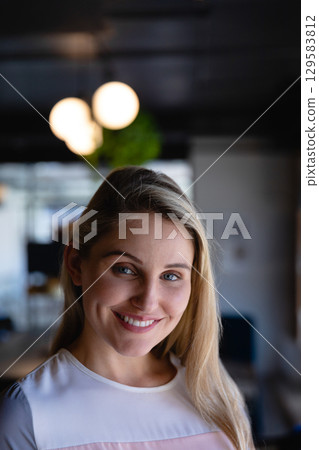 Businesswoman wearing white blouse standing at desk smiling in office under globe lights near plant Businesswoman wearing white blouse standing at desk smiling in office under globe lights near plant 129583812