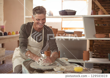 Senior potter wearing apron shaping wet clay vessel on potter's wheel in clay studio with shelving 129583819