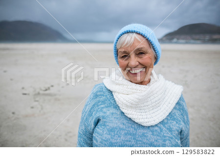 Senior woman smiling on sandy beach at low tide wearing blue beanie, white scarf, copy space 129583822