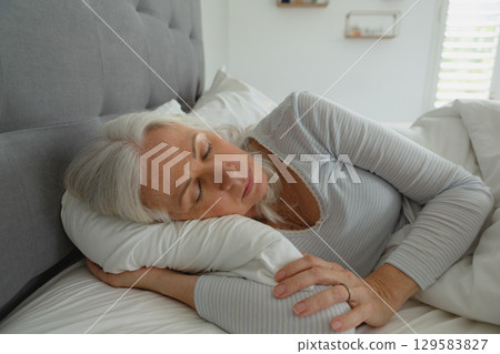 Mature woman lying on side in bedroom showing white pillow comforter grey headboard blinds Mature woman lying on side in bedroom showing white pillow comforter grey headboard blinds 129583827