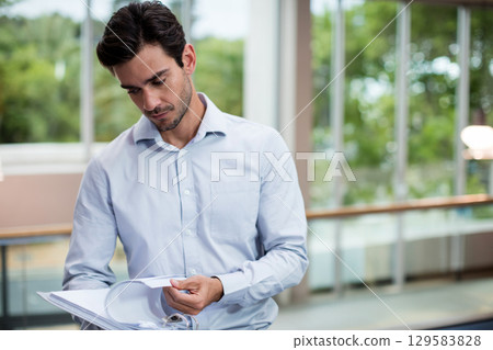 Mid adult man standing in lobby flipping through white three-ring binder pages by glass railing Mid adult man standing in lobby flipping through white three-ring binder pages by glass railing 129583828