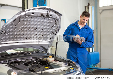 Male mechanic logging engine bay data with clipboard and pen near hydraulic lift posts, copy space 129583830