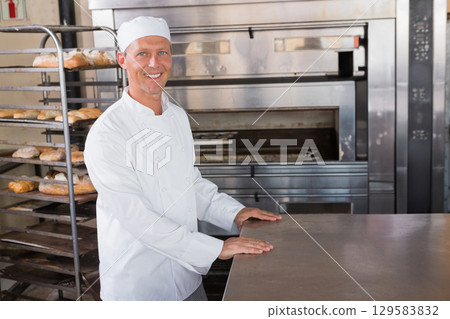 Mature male baker arranging bread loaves on metal rack near stainless steel countertop and ovens 129583832