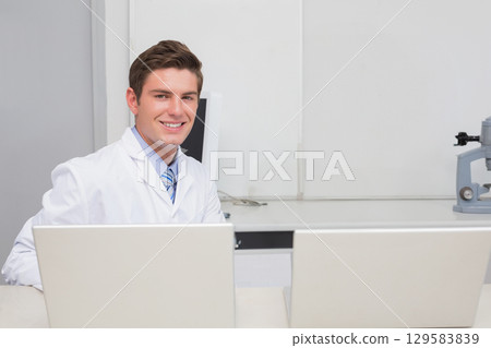 Male optometrist wearing white coat sitting at desk in exam room with two laptops and lensometer 129583839
