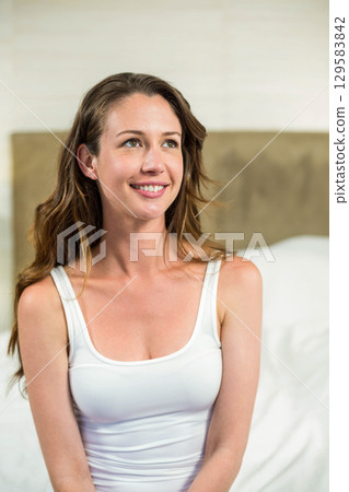 Woman in twenties sitting on bed in bedroom wearing white tank top with headboard and pillows 129583842
