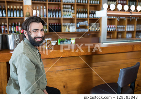 Smiling bearded man in twenties sitting at bar counter holding beer glass metal tap, copy space 129583848