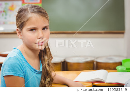Girl child wearing blue t-shirt writing in notebook at wooden desk in classroom with chalkboard 129583849
