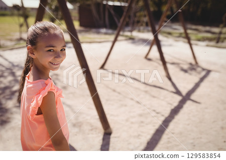 School-age girl standing on sandy playground at suburban park with wooden swing frames, copy space 129583854