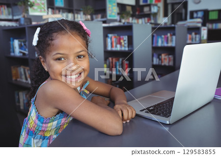 African American girl sitting at table in library using silver laptop by bookshelves, copy space 129583855
