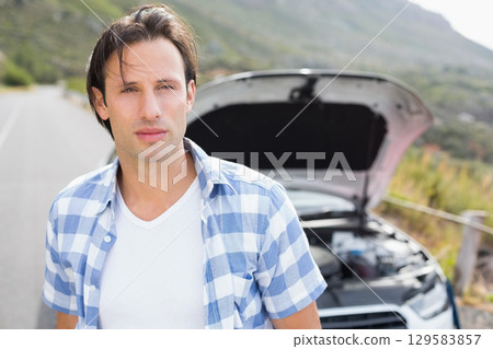 Middle-aged man standing beside vehicle with open hood on rural road examining engine components 129583857