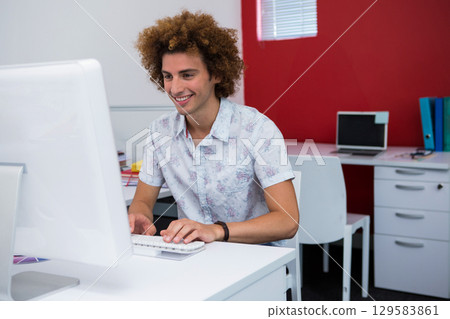 Man in late twenties wearing short-sleeve shirt typing on keyboard at desk in office with binders 129583861
