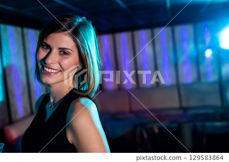 Woman smiling in black sleeveless top and heart pendant while standing in nightclub, copy space 129583864