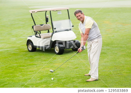 Senior African American man preparing to swing club at golf ball on fairway beside golf cart 129583869