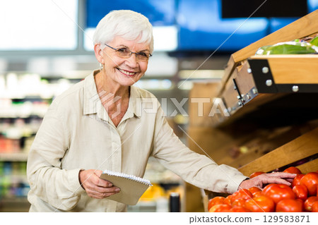 Senior woman inspecting ripe tomatoes on wooden produce bin holding spiral notebook in supermarket Senior woman inspecting ripe tomatoes on wooden produce bin holding spiral notebook in supermarket 129583871