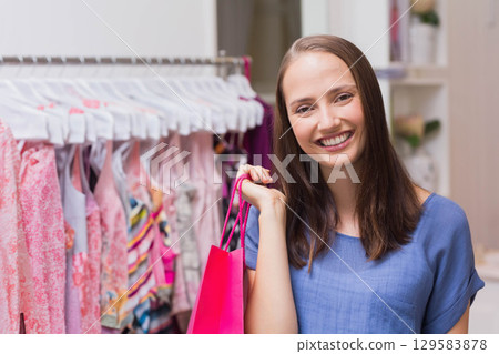 Woman standing in boutique store holding pink shopping bag beside pastel dress rack, copy space Woman standing in boutique store holding pink shopping bag beside pastel dress rack, copy space 129583878