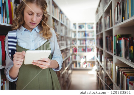 Child girl leaning on left bookshelves using tablet in library with striped backpack, copy space 129583891