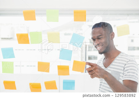 African American man smiling, pointing at sticky note on partition by office windows, copy space 129583892