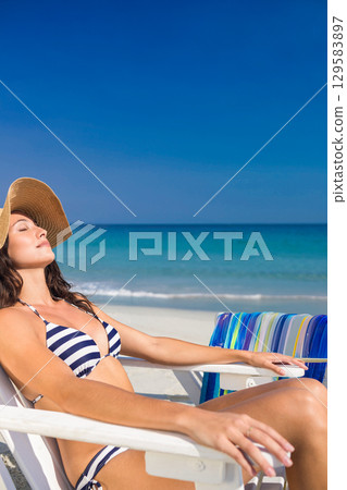 Woman reclining on lounge chair at shoreline wearing striped bikini with straw sun hat, copy space 129583897