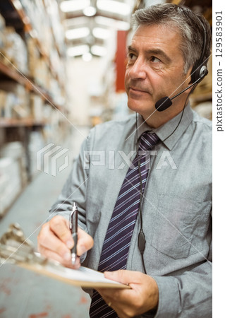 Man wearing tie inspecting boxes in warehouse aisle holding clipboard and headset, copy space Man wearing tie inspecting boxes in warehouse aisle holding clipboard and headset, copy space 129583901
