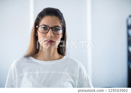 Woman standing against off-white wall in room wearing tortoiseshell eyeglasses and white shirt Woman standing against off-white wall in room wearing tortoiseshell eyeglasses and white shirt 129583902