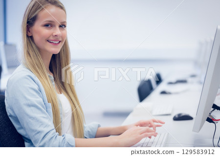Teenage girl smiling while typing at computer lab desk with keyboard and monitor, copy space 129583912