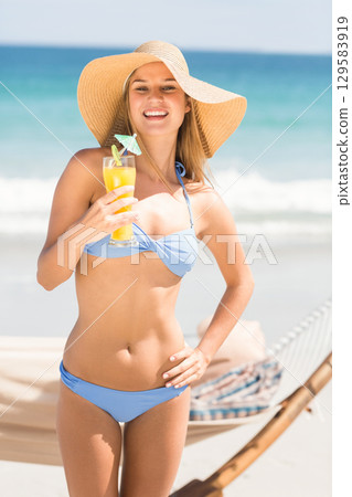 Female tourist holding glass of orange juice in swimsuit and straw hat with hammock on beach 129583919