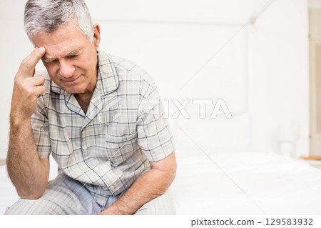 Senior man pressing temple while on bed edge in bedroom with lamp, plaid pajamas, copy space Senior man pressing temple while on bed edge in bedroom with lamp, plaid pajamas, copy space 129583932
