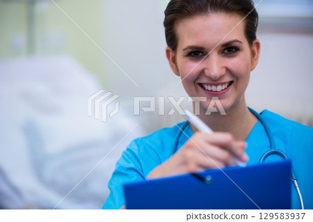 Female nurse smiling filling out chart by bed in ward with clipboard, stethoscope, copy space 129583937