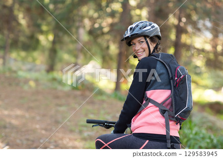 Female cyclist sitting on mountain bike on forest trail wearing helmet with backpack, copy space 129583945
