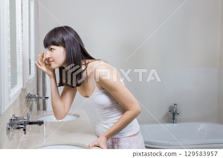 Woman leaning over marble vanity mirror adjusting skincare under left eye in bathroom, copy space Woman leaning over marble vanity mirror adjusting skincare under left eye in bathroom, copy space 129583957