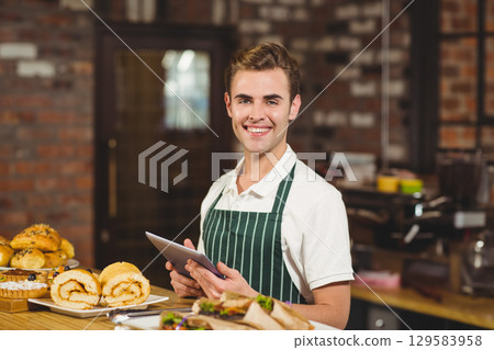 Mid-adult man in striped apron holding tablet behind counter in bakery near bread rolls Mid-adult man in striped apron holding tablet behind counter in bakery near bread rolls 129583958