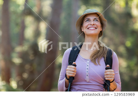 Female hiker standing on forest trail holding poles wearing lavender top bucket hat and backpack Female hiker standing on forest trail holding poles wearing lavender top bucket hat and backpack 129583969