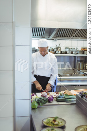 Male chef slicing vegetables on wooden cutting board in professional kitchen near stainless pots 129583970