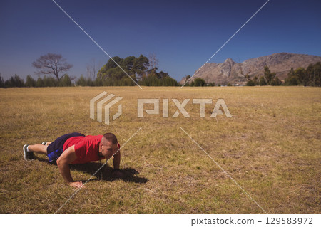 Man performing push-ups on grass wearing red shirt running shoes, whistle lanyard, copy space 129583972