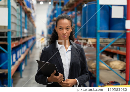 African American businesswoman standing in warehouse aisle reviewing clipboard near metal racks 129583980