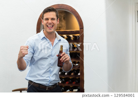 Man standing in wine room holding beer bottle and raising fist by wine rack, copy space 129584001