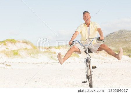 Middle-aged man riding beach cruiser bicycle along beach dunes while kicking legs up and laughing 129584017