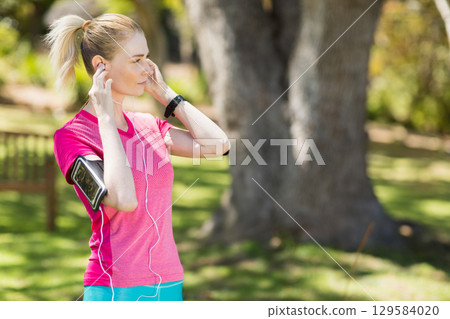 Woman in sportswear standing in park adjusting earphones with phone armband and tracker, copy space 129584020