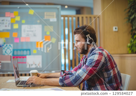 Man wearing plaid shirt and headphones working on laptop at office near sticky notes, copy space 129584027