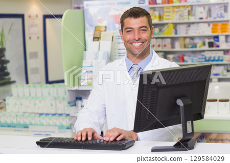 Male pharmacist wearing lab coat standing behind counter at pharmacy with monitor and keyboard Male pharmacist wearing lab coat standing behind counter at pharmacy with monitor and keyboard 129584029