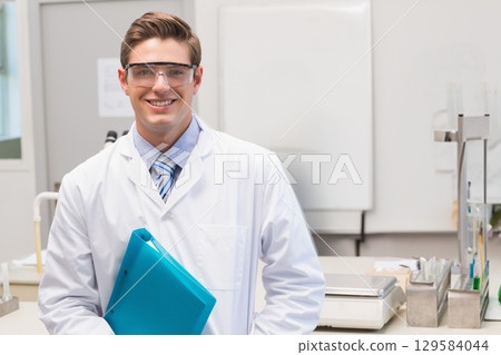 Male scientist standing in laboratory wearing white lab coat and goggles while holding blue folder Male scientist standing in laboratory wearing white lab coat and goggles while holding blue folder 129584044