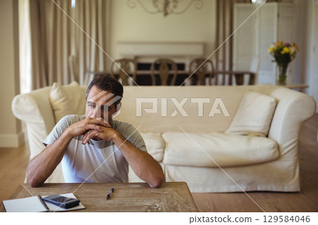 Middle-aged man sitting at wooden coffee table beside sofa in living room using calculator and pen Middle-aged man sitting at wooden coffee table beside sofa in living room using calculator and pen 129584046
