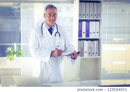 Male physician standing in clinic writing on clipboard wearing stethoscope near laptop, binders 129584051