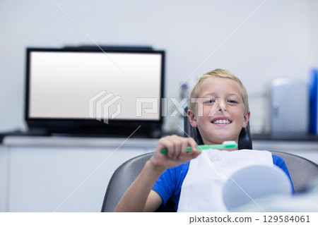 Smiling boy reclining in dental chair holding toothbrush near dental mirror at clinic, copy space Smiling boy reclining in dental chair holding toothbrush near dental mirror at clinic, copy space 129584061