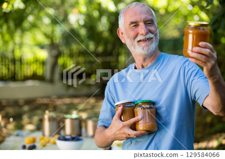 Senior man holding amber preserve jars and placing fruit bowls on wooden table in backyard garden Senior man holding amber preserve jars and placing fruit bowls on wooden table in backyard garden 129584066